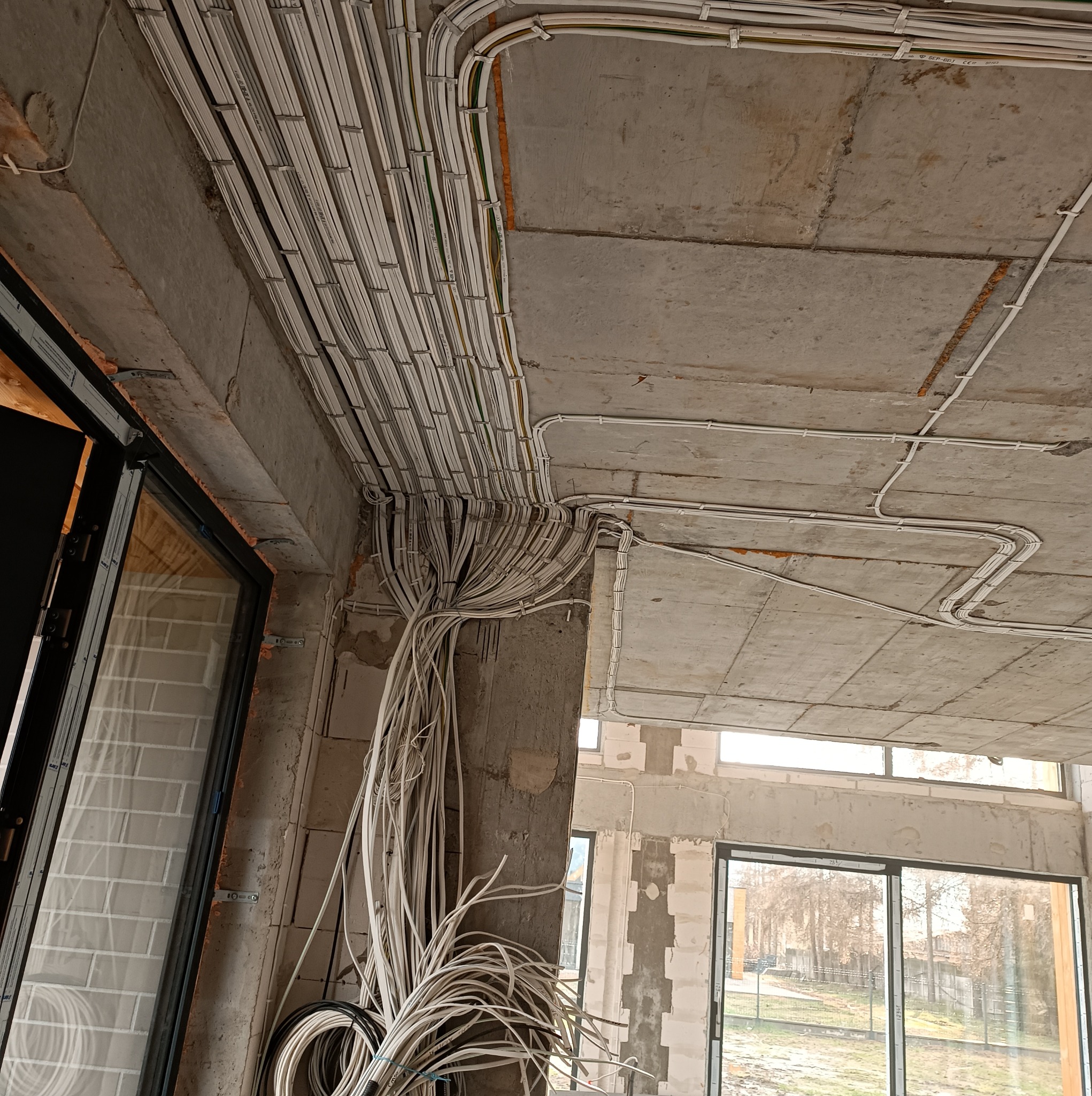 Białe kable elektryczne ułożone w rurkach na surowym betonowym suficie w trakcie budowy, widoczne okna i surowy stan wykończenia.