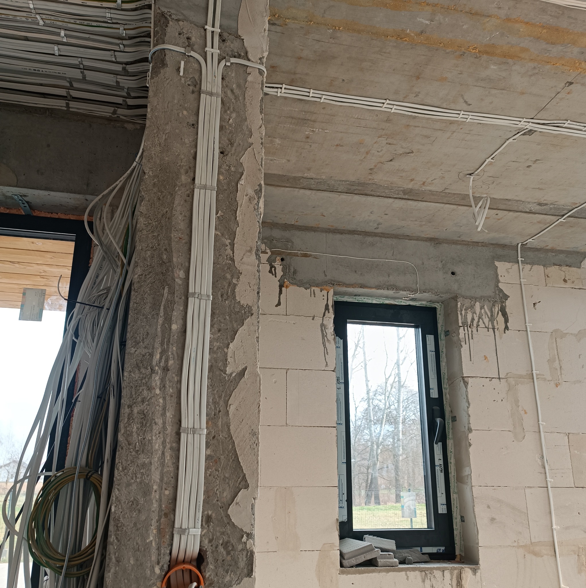 Surowy stan instalacji elektrycznej w nowym budynku: wiązki przewodów na ścianie z betonu komórkowego, kable prowadzone po suficie, widoczne okno z widokiem na drzewa w tle.