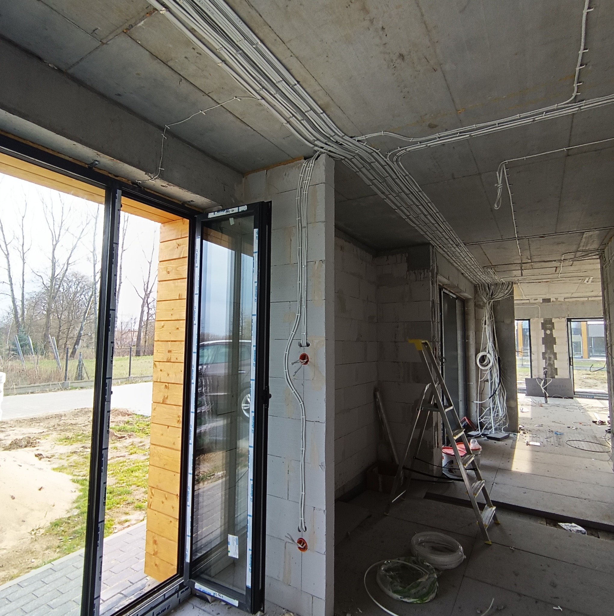 Wnętrze w stanie surowym w Piasecznie z widocznymi przewodami elektrycznymi prowadzonymi po suficie i ścianach, otwór na gniazdko elektryczne w ścianie, drabina oparta o ścianę, duże okno...