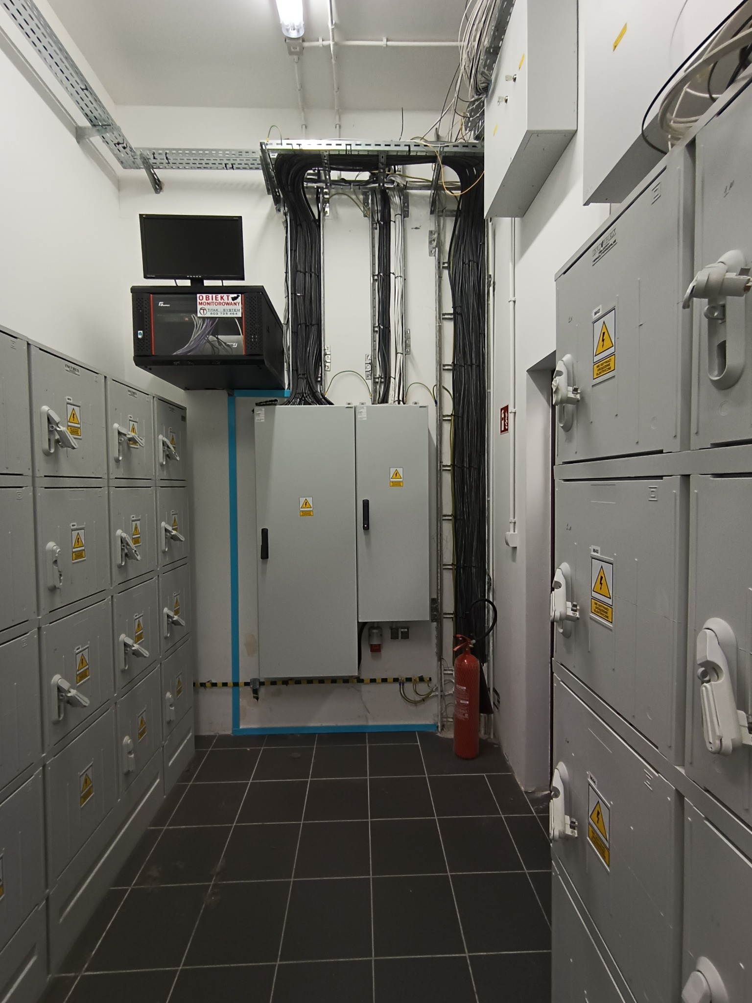 Wnętrze pomieszczenia z szafami elektrycznymi, rozdzielnicą, kablami prowadzonymi w korytach oraz gaśnicą w rogu, monitor i serwer na szafkach po lewej stronie.