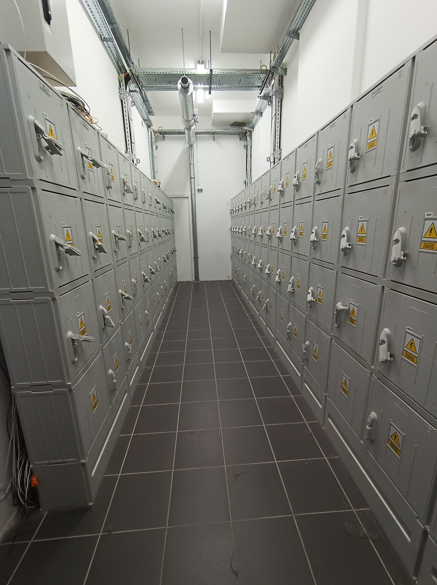 Rząd szarych szaf elektrycznych z żółtymi znakami ostrzegawczymi w pomieszczeniu z ciemnymi płytkami podłogowymi i metalowymi korytami kablowymi pod sufitem.