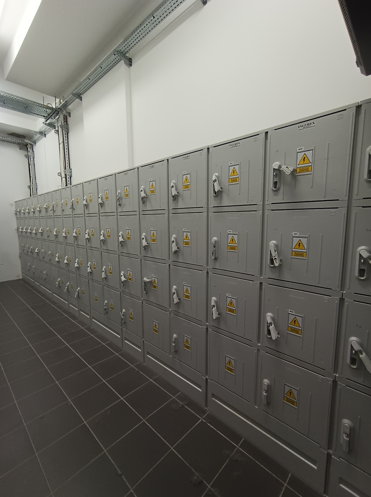 Rząd szarych szaf elektrycznych z ostrzeżeniami o wysokim napięciu w pomieszczeniu z szarą podłogą z płytek i metalowymi korytami kablowymi pod sufitem.