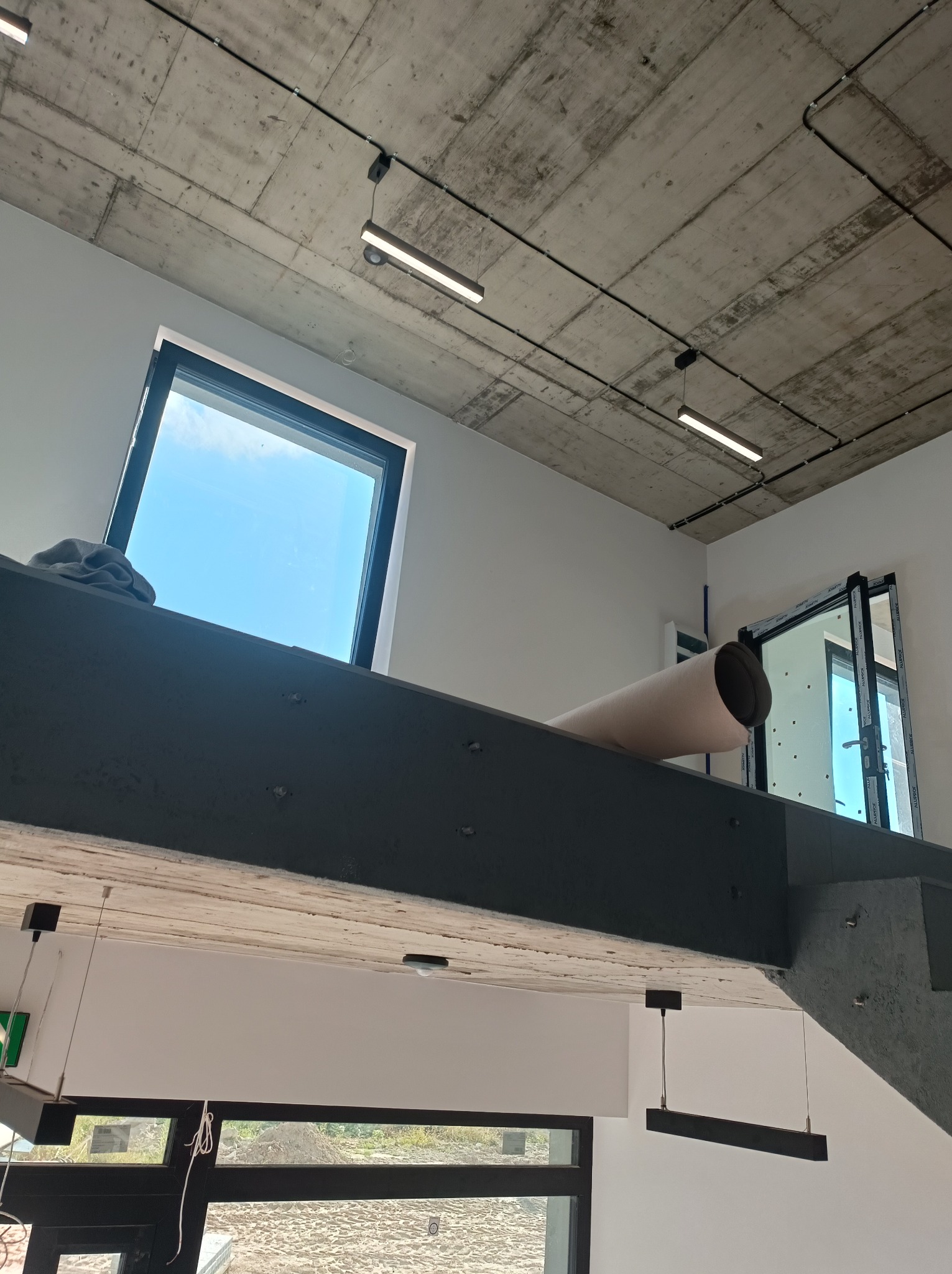 Surowy betonowy sufit z widocznymi przewodami elektrycznymi i lampami LED, widok z dołu na antresolę z oknem i rolką materiału budowlanego