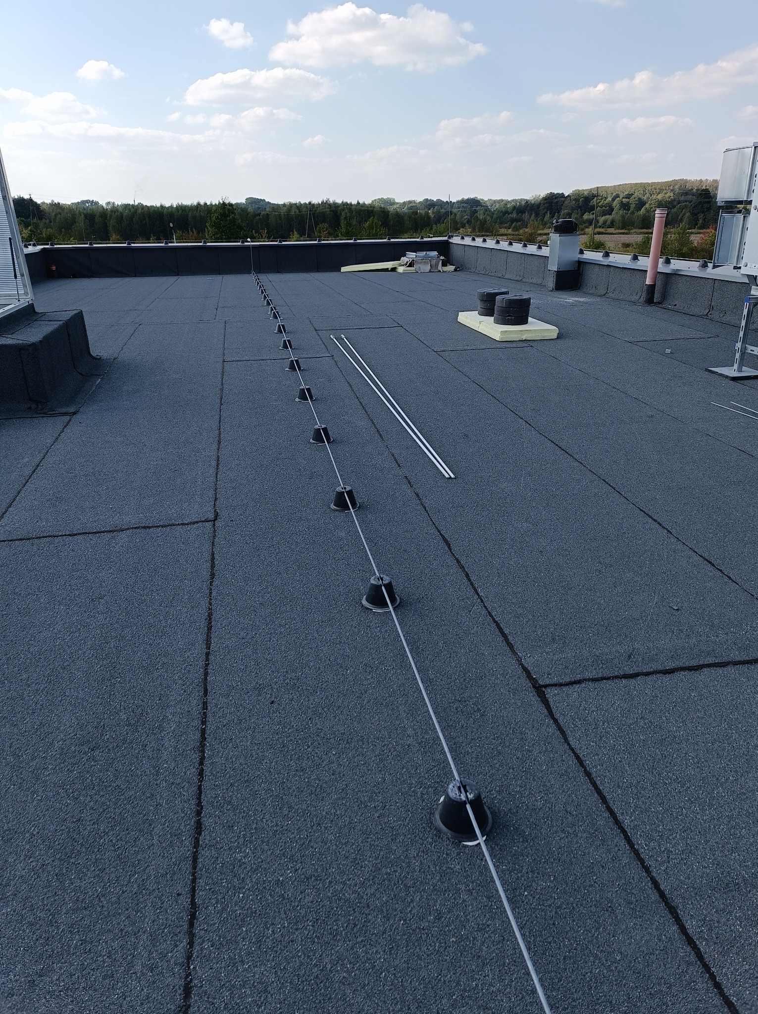 Płaski dach pokryty papą z zamontowanym systemem odgromowym, widoczne czarne uchwyty i linka, w tle zieleń drzew i błękitne niebo.