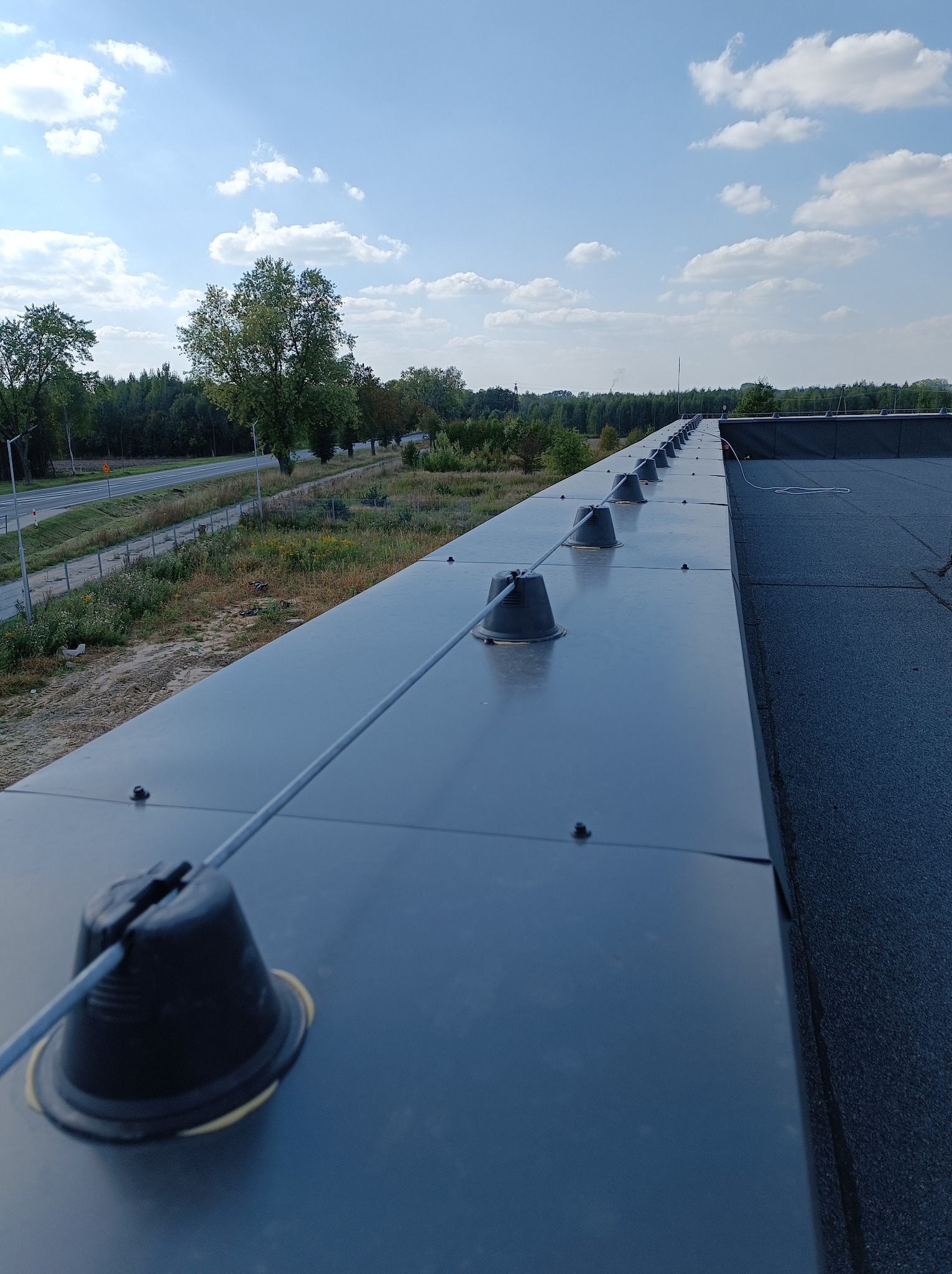 Widok z góry na zainstalowany system odgromowy na dachu budynku, z widocznymi uchwytami i przewodami, otoczony zielenią i błękitnym niebem.