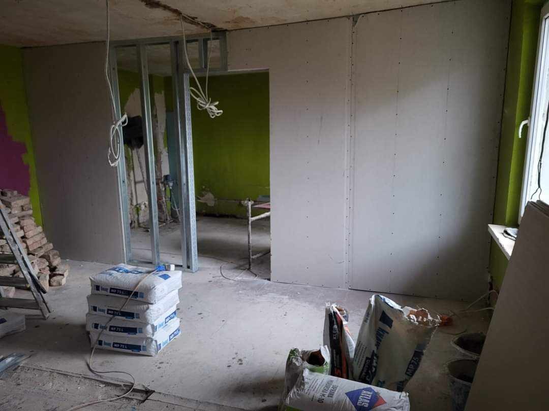 Wnętrze w trakcie prac: zamontowane płyty kartonowo-gipsowe na ścianie, stalowa konstrukcja sufitowa, worki z zaprawą Atlas i MP75 na podłodze, widoczne kable i drabina.