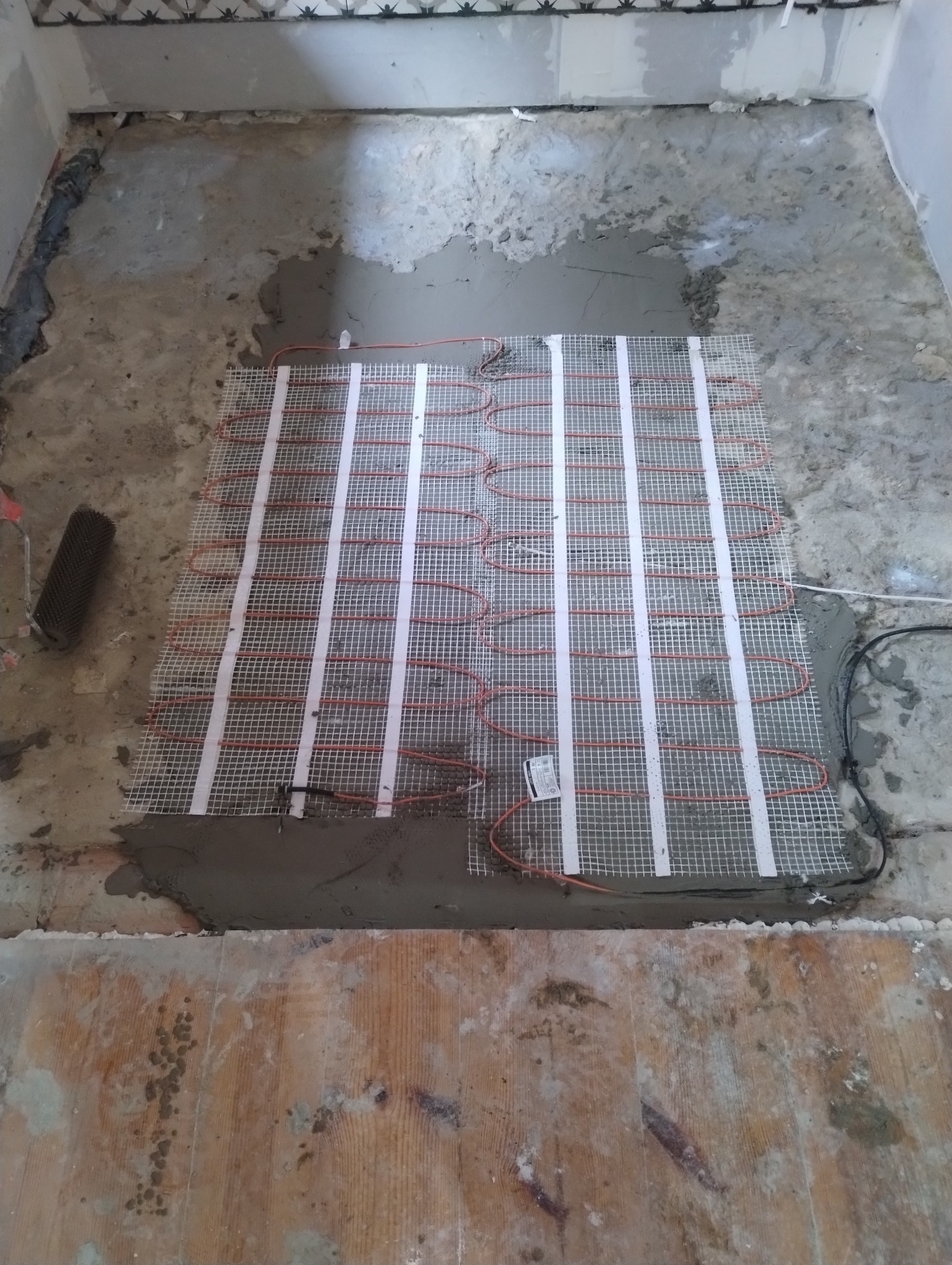 Mata grzewcza podłogowa rozłożona na wylewce, przygotowana do zalania klejem, widoczny fragment drewnianej podłogi i narzędzie do rozprowadzania kleju.