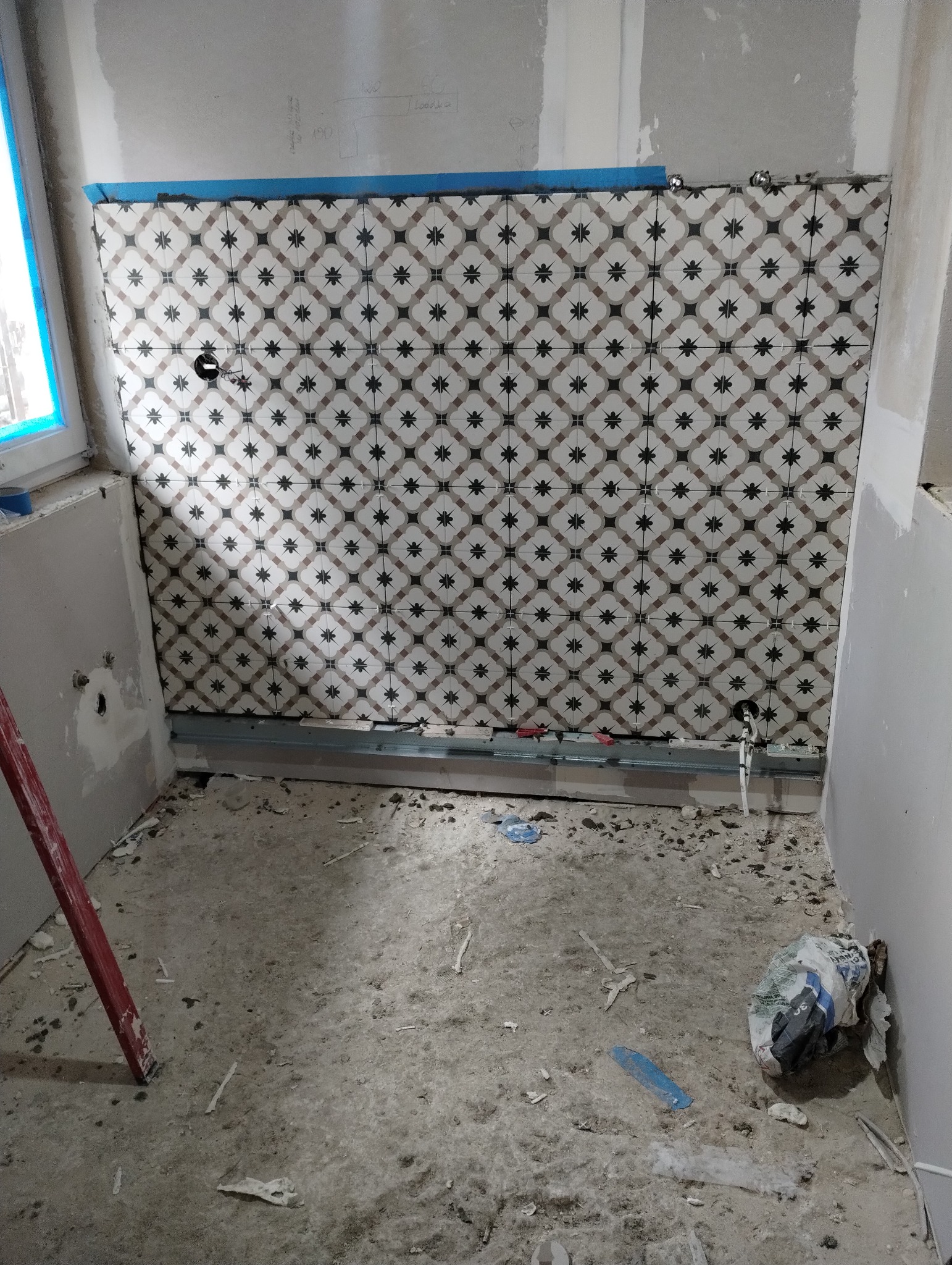 Ściana w łazience w trakcie układania płytek z dekoracyjnym wzorem, widoczne nieotynkowane ściany, fragment okna z niebieską folią ochronną, metalowy profil pod płytkami i resztki zaprawy na podłodze.
