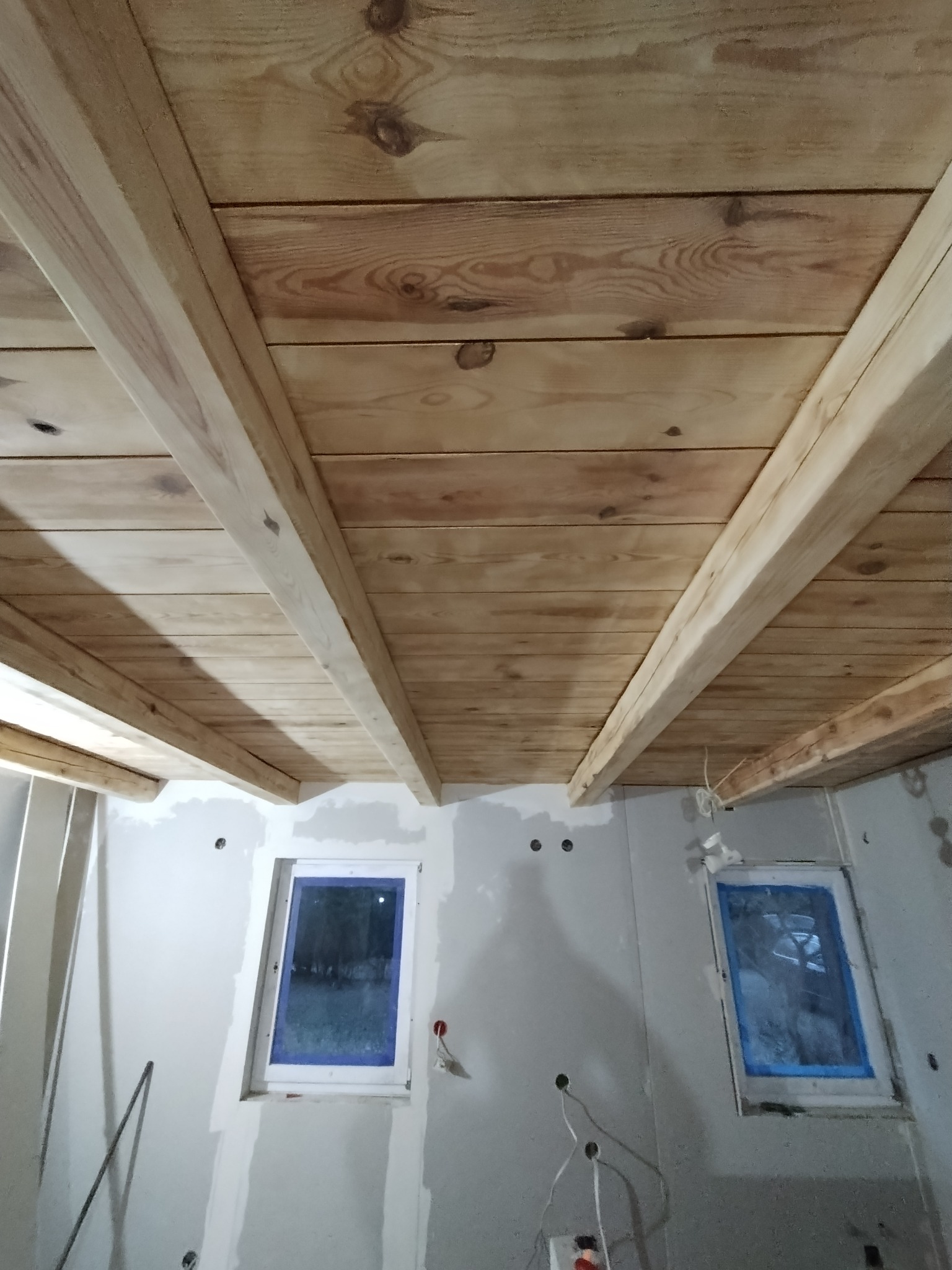 Drewniany strop z belkami na poddaszu w trakcie prac wykończeniowych, widoczne okna zabezpieczone folią i ściany przygotowane do dalszych prac.