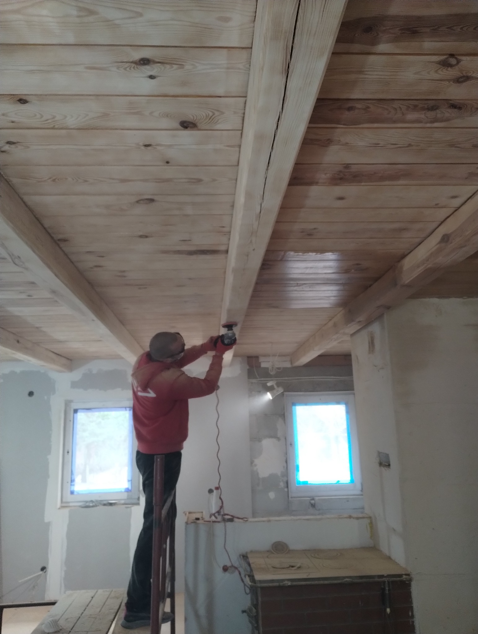 Pracownik w czerwonej bluzie szlifuje drewniany sufit z widocznymi belkami za pomocą szlifierki elektrycznej, stojąc na prowizorycznym rusztowaniu w jasnym, remontowanym pomieszczeniu.