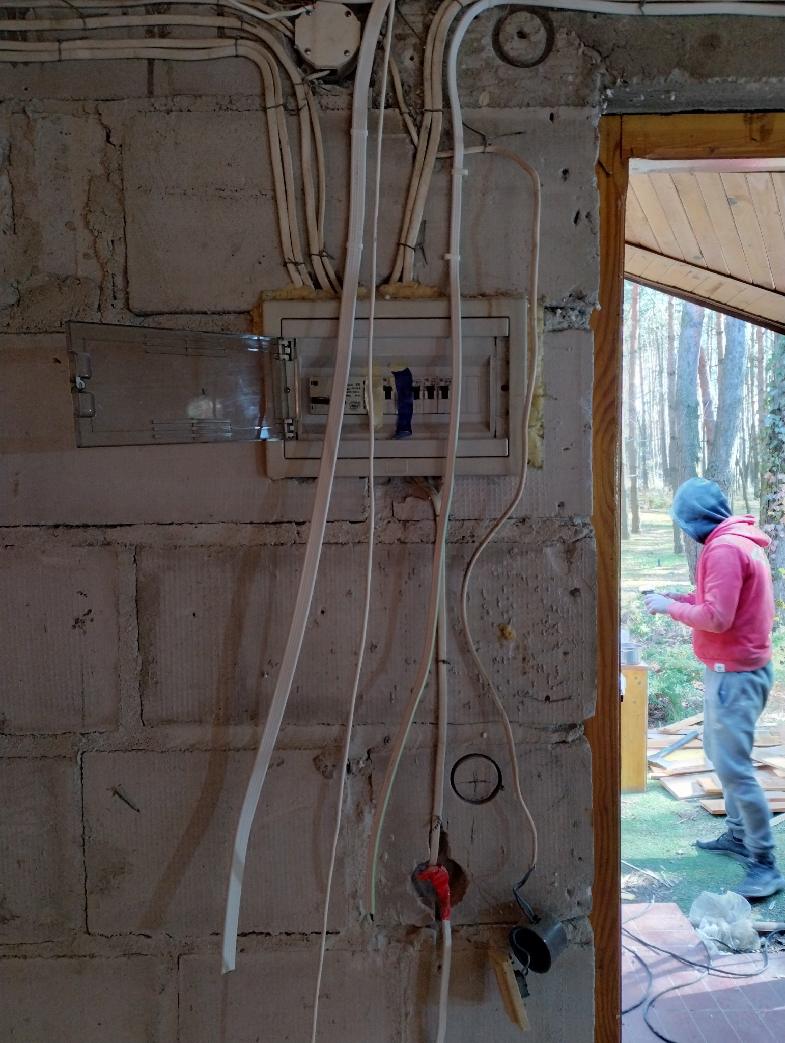 Rozdzielnia elektryczna z otwartą klapką na surowej ścianie z bloków, widoczne kable i otwory, w tle osoba w bluzie przy oknie.