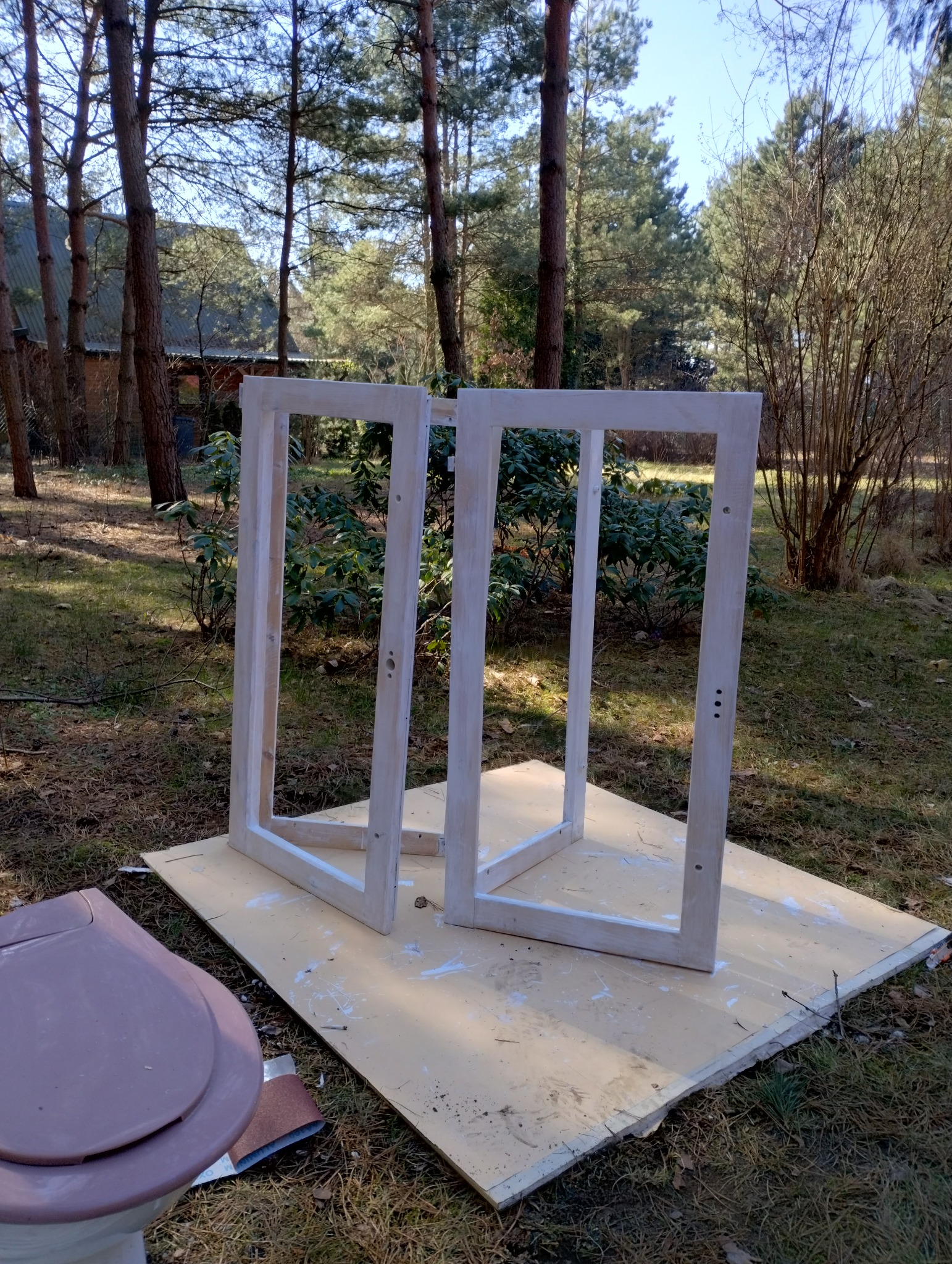 Dwie drewniane ramy pomalowane na biało, ustawione na płycie z resztkami farby, na tle ogrodu z drzewami iglastymi i liściastymi; w pobliżu leży pokrywa toalety i papier ścierny.