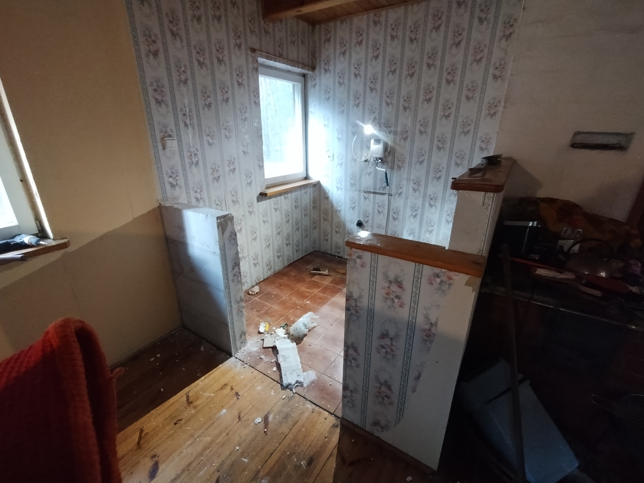Demontaż ścianki działowej w pokoju z tapetą w kwiaty i drewnianą podłogą, widoczne pozostałości gruzu i narzędzia.