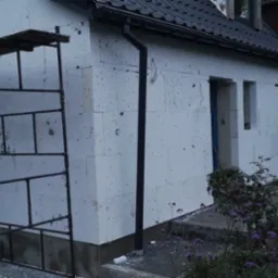 Ocieplanie ścian zewnętrznych budynku styropianem, widoczne rusztowanie i poziomica, okna zabezpieczone folią, przygotowane otwory okienne.