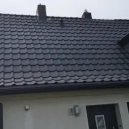 Nowoczesny dom z ciemnoszarą dachówką ceramiczną, widoczne okno dachowe z folią ochronną, rynna i fragment elewacji z numerem domu.