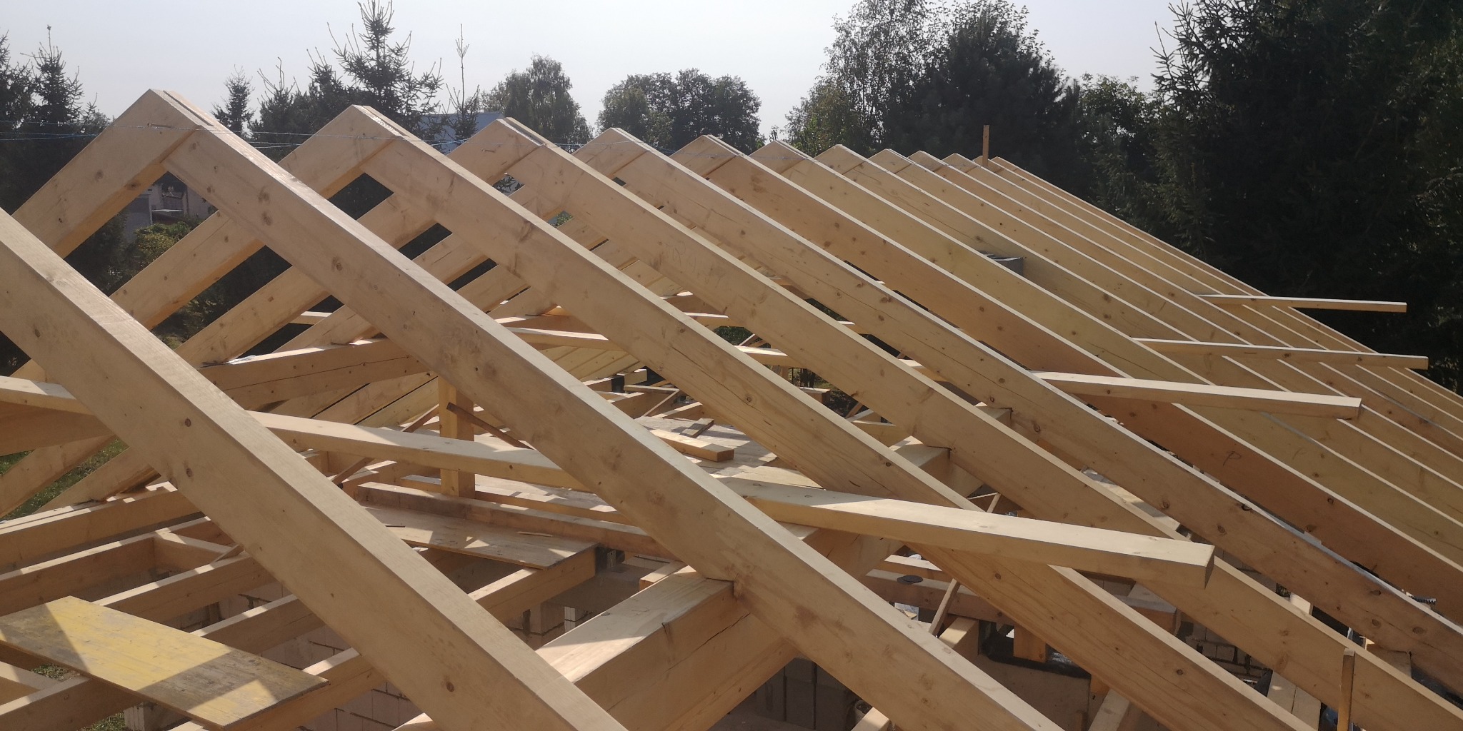 Surowa drewniana konstrukcja więźby dachowej w trakcie montażu, widok z dołu na tle lasu.
