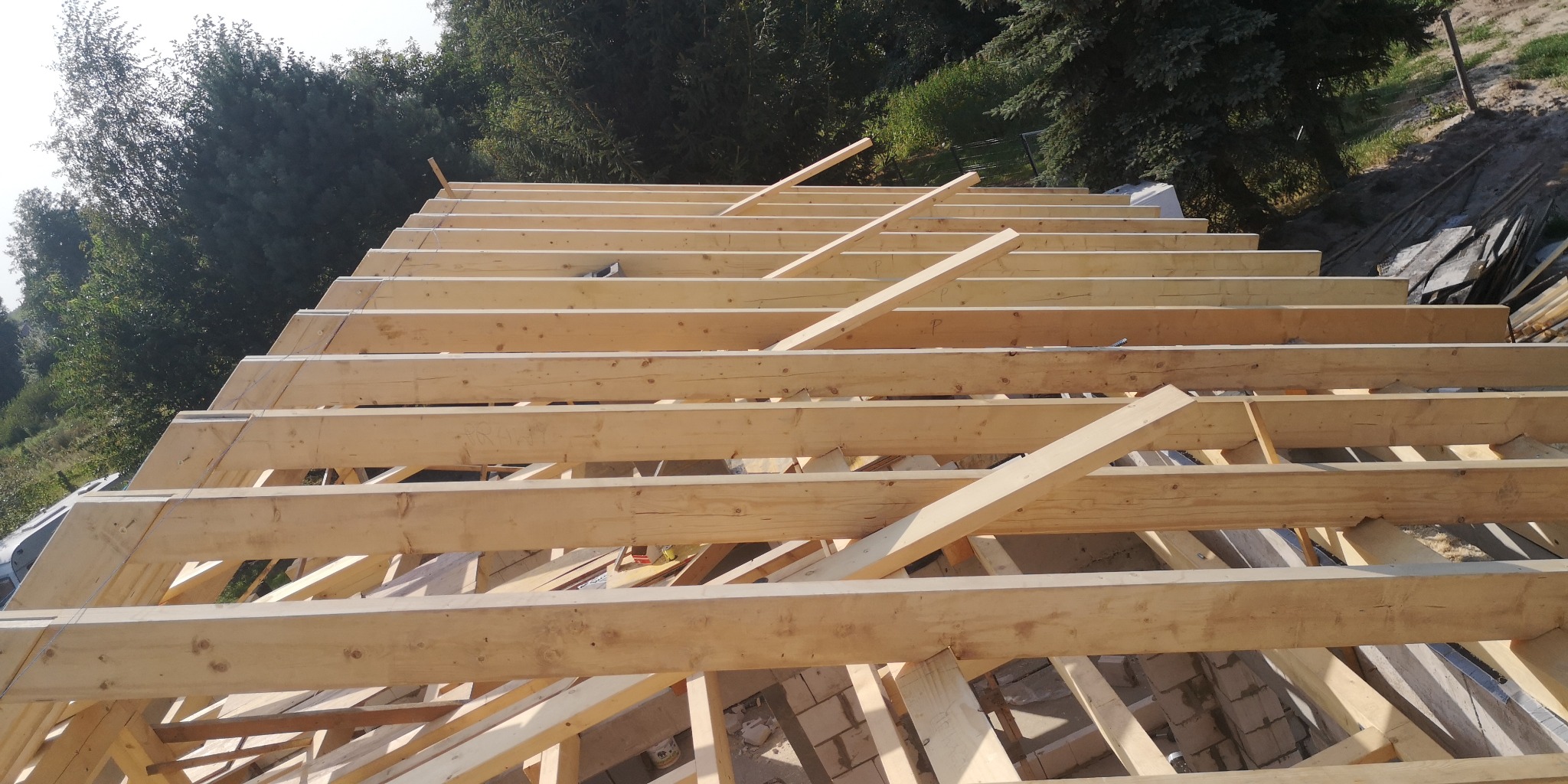 W trakcie budowy drewniana konstrukcja dachu z widocznymi belkami i krokwiami, w tle zieleń drzew.