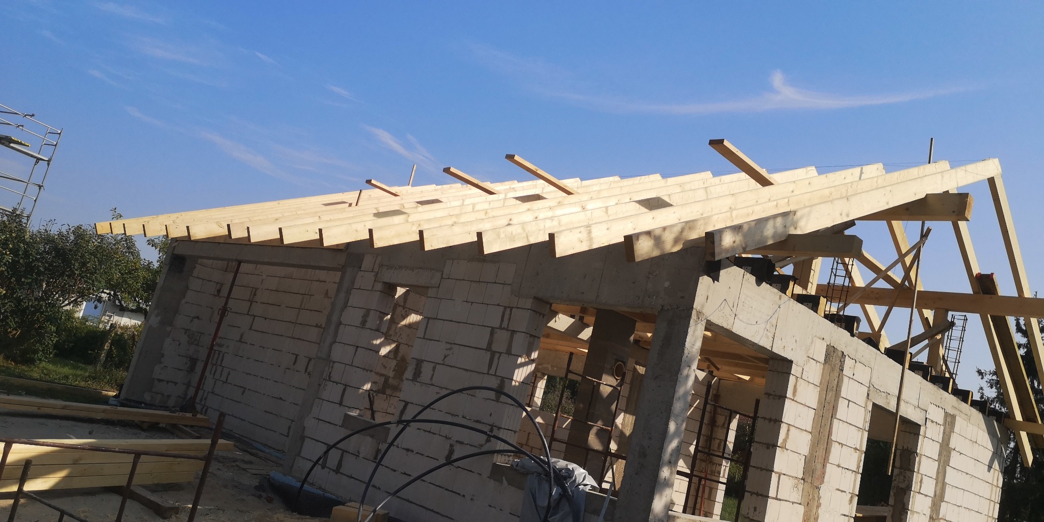 Wznoszenie drewnianej więźby dachowej na murowanym budynku w stanie surowym otwartym, widoczne elementy konstrukcyjne dachu i fragment rusztowania.