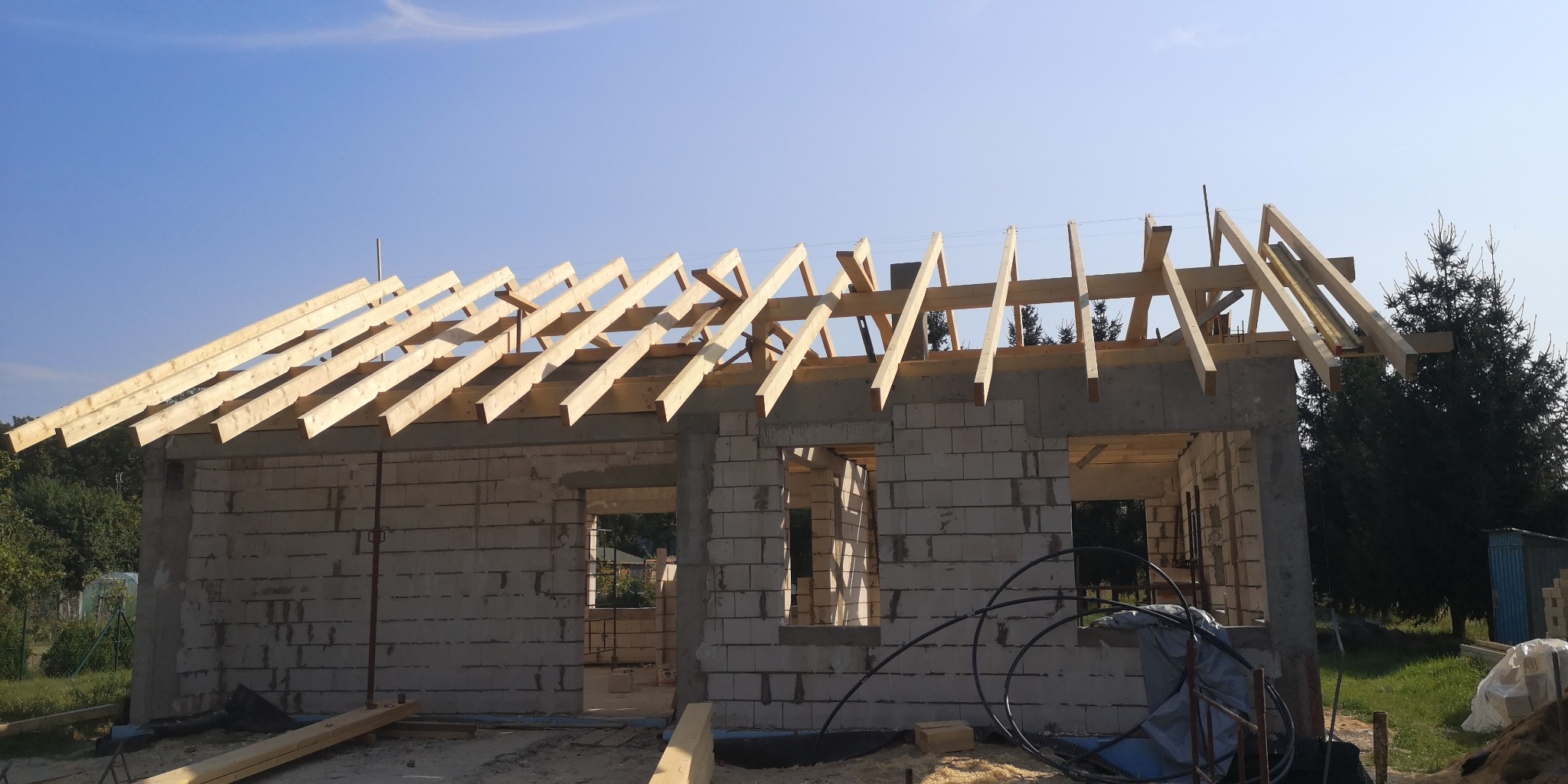Surowy stan budynku z betonu komórkowego z częściowo wykonaną drewnianą konstrukcją dachu, widoczne wiązary krokwiowe pod kątem na tle błękitnego nieba.