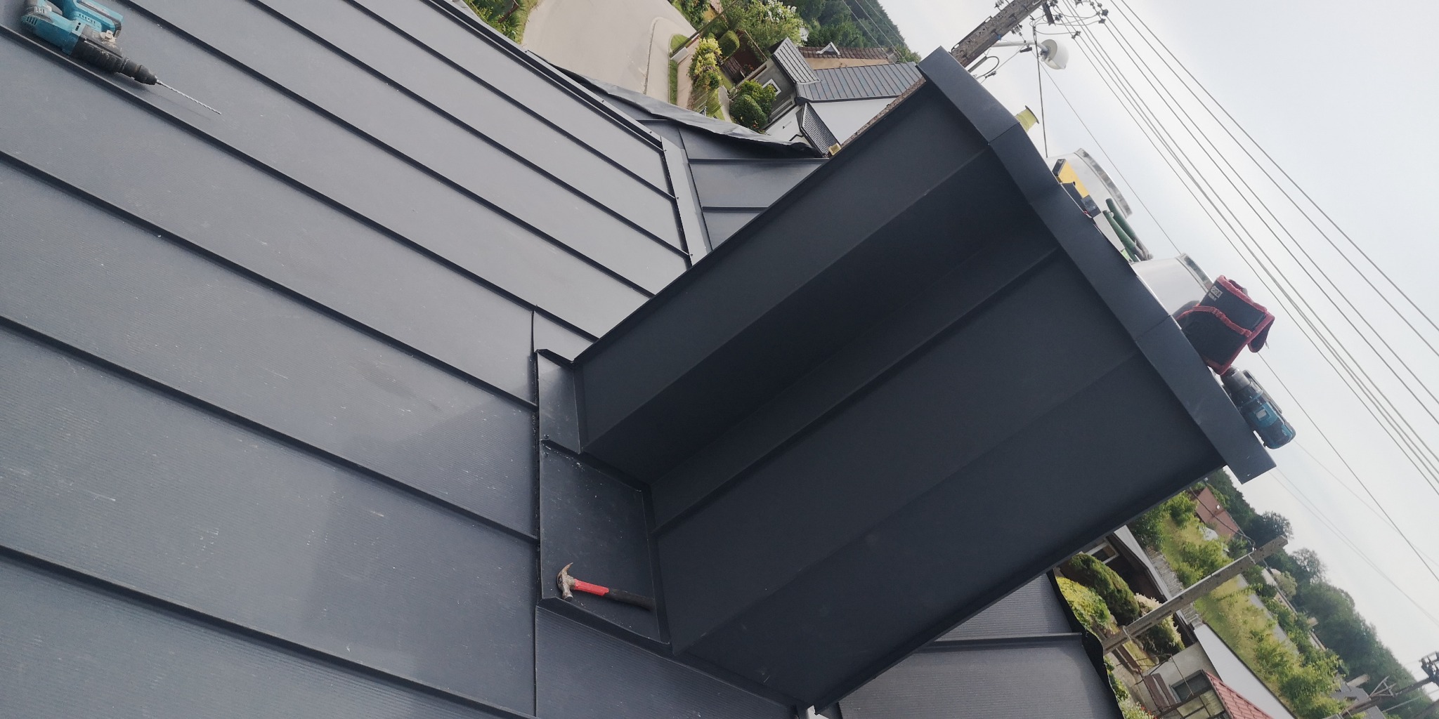 Nowoczesne, grafitowe pokrycie dachowe z blachy na rąbek stojący, z narzędziami: wkrętarką i młotkiem, widoczne elementy obróbek blacharskich przy lukarnie.