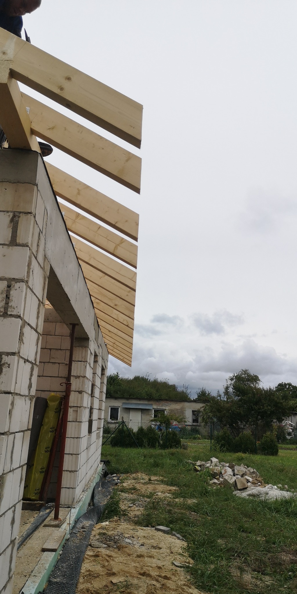 Wznoszenie więźby dachowej z drewnianych belek na budynku z białego bloczka, widok z dołu na pochmurne niebo i częściowo zarośnięte budynki w tle.