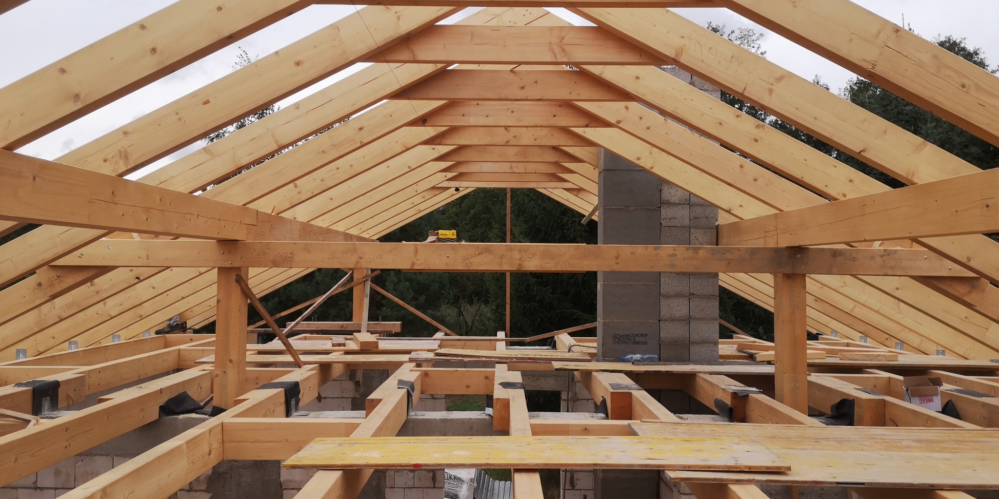 Surowa konstrukcja dachu z drewna z widocznym kominem z pustaków, widok od wewnątrz, w tle zieleń drzew.