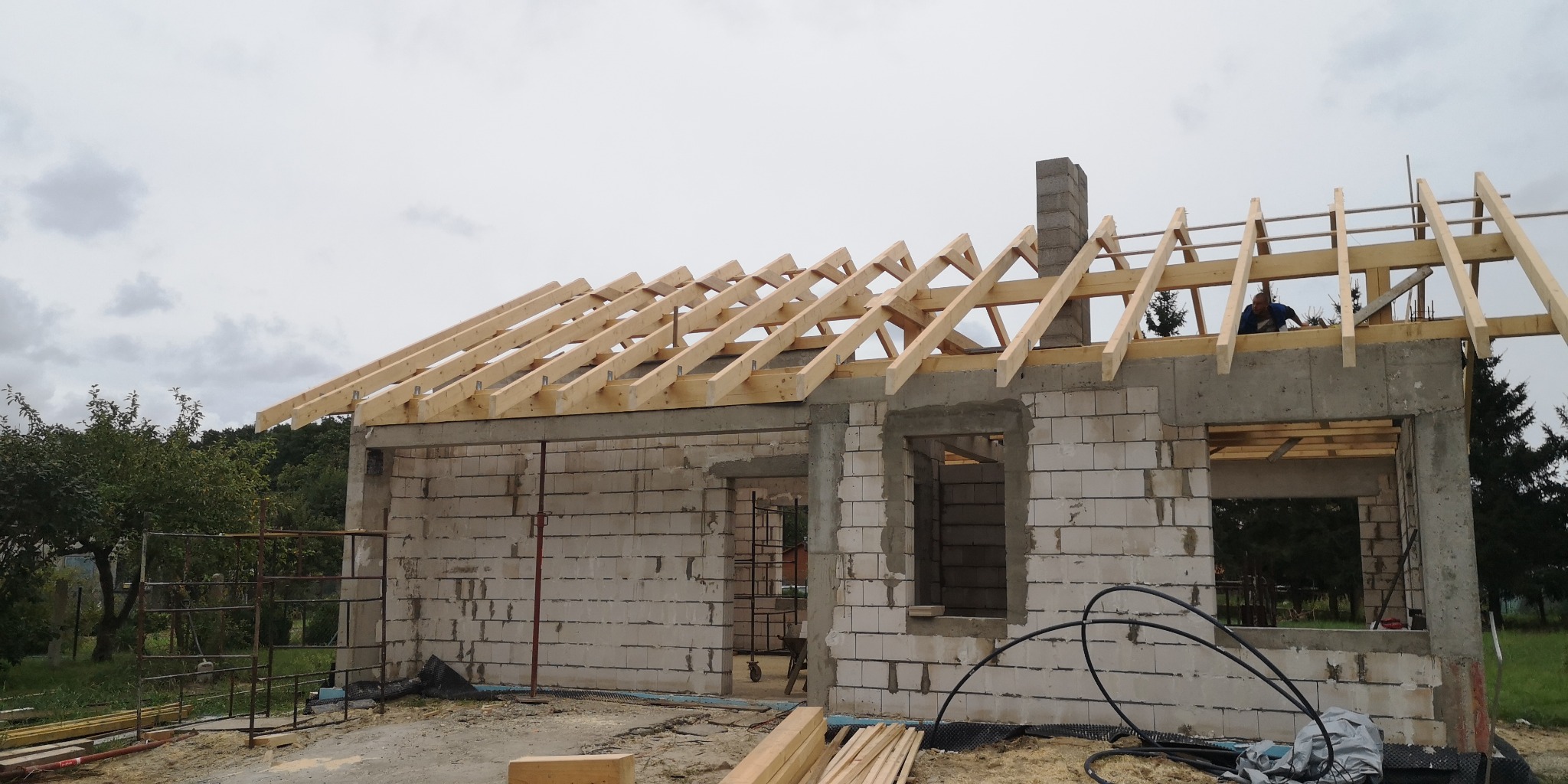 Wznoszenie drewnianej konstrukcji dachu na murowanym domu jednorodzinnym w trakcie budowy, widoczny komin i pracownik na dachu.