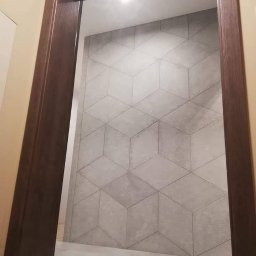 Roman Dotsenko - Ściana wyłożona szarymi płytkami w geometryczny wzór 3D, widoczna z perspektywy wejścia z ciemną, drewnianą futryną.