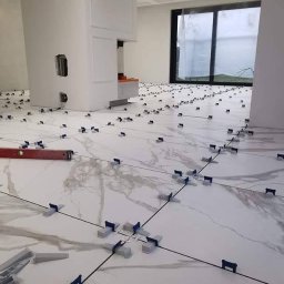 Roman Dotsenko - Układanie płytek podłogowych imitujących marmur w przestronnym, jasnym wnętrzu; widoczne krzyżyki dystansowe i poziomica laserowa, w tle okno i dekoracyjny sufit.