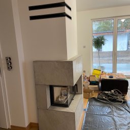 Roman Dotsenko - Montaż obudowy kominka z płyt kamiennych, widoczne czarne listwy dekoracyjne na ścianie, podłoga zabezpieczona folią, w tle okno tarasowe.