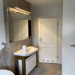 Roman Dotsenko - Łazienka w trakcie prac wykończeniowych: umywalka z szafką, lustro z oświetleniem LED, płytki imitujące kamień na ścianie, białe drzwi z widocznymi otworami montażowymi w dolnej części.