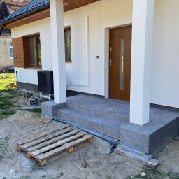 Roman Dotsenko - Wykończone schody wejściowe do domu z szarych płytek, drewniane drzwi wejściowe, białe kolumny i elewacja, widoczna paleta i materiały budowlane na zewnątrz.