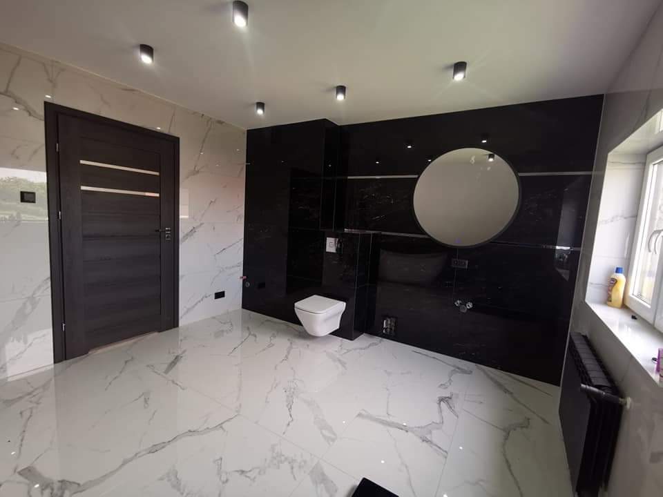 Nowoczesna łazienka z białymi marmurowymi płytkami na podłodze i ścianach, kontrastującymi z czarną ścianą z okrągłym lustrem i podwieszaną toaletą, oświetlona punktowymi lampami sufitowymi