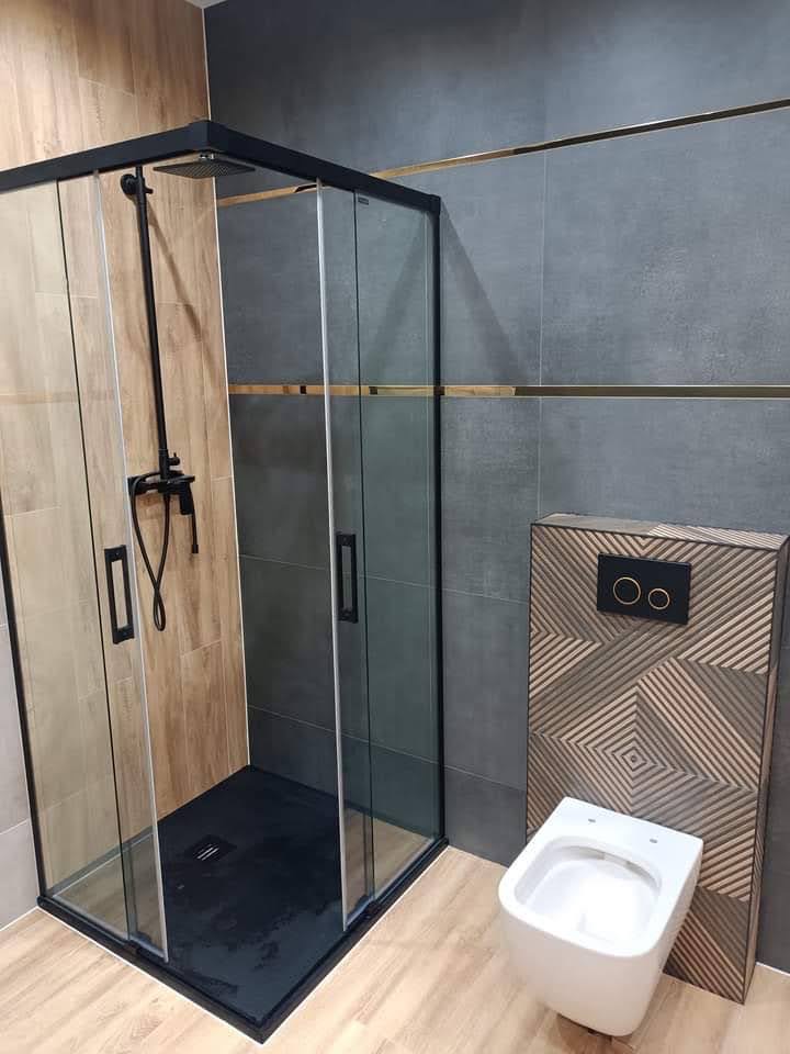 Nowoczesna łazienka z kabiną prysznicową typu walk-in z czarnymi profilami, podwieszaną miską WC z dekoracyjną obudową i szarymi płytkami z złotymi akcentami.