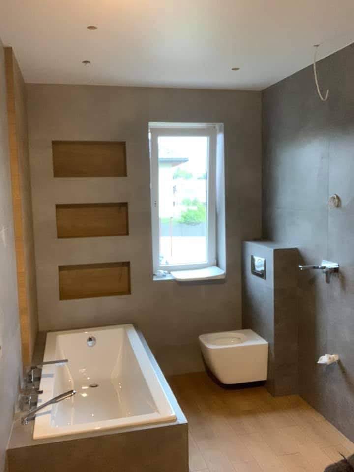 Nowoczesna łazienka z wanną obudowaną szarą płytką, podwieszaną toaletą i drewnianą podłogą; widoczne trzy wnęki w ścianie wyłożone drewnem; okno z widokiem na zieleń.