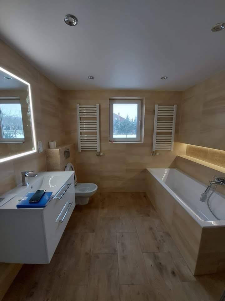 Nowoczesna łazienka z beżowymi płytkami imitującymi drewno, podświetlanym lustrem, grzejnikami drabinkowymi i wanną w zabudowie, widok z góry na telefon leżący na blacie szafki umywalkowej.