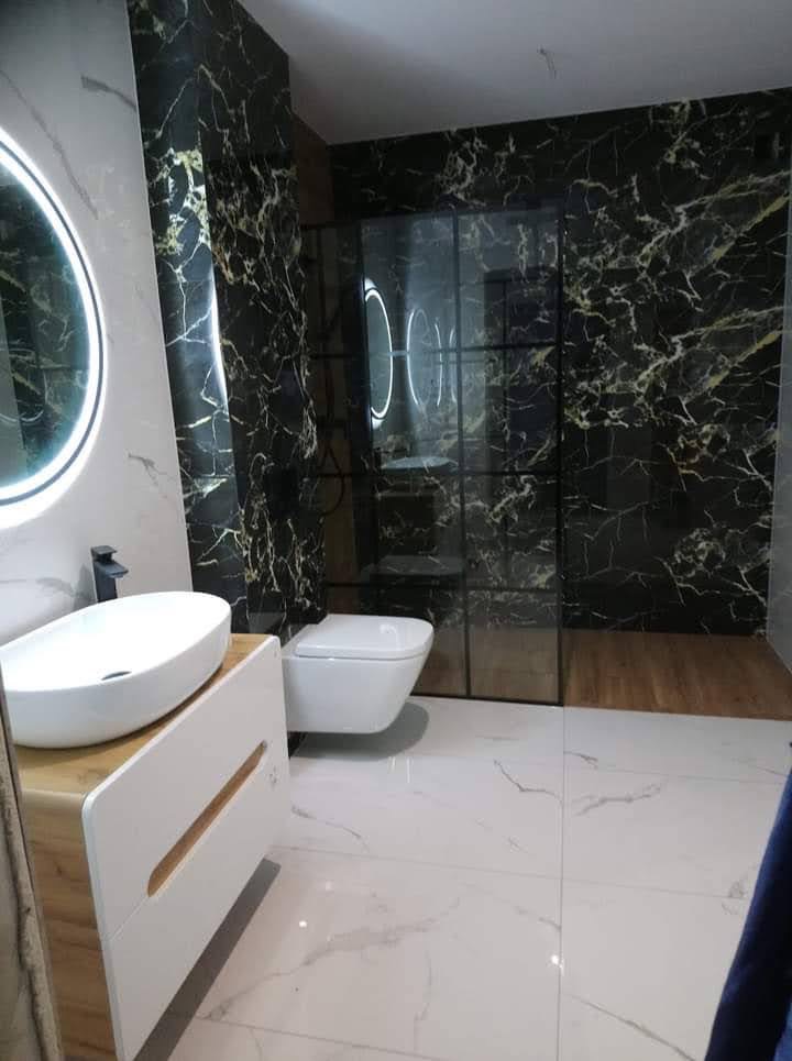 Nowoczesna łazienka z czarnymi płytkami imitującymi marmur, szklaną kabiną prysznicową w stylu loft, podwieszaną toaletą i umywalką nablatową na szafce z drewnianymi akcentami, oświetlona okrągłym...