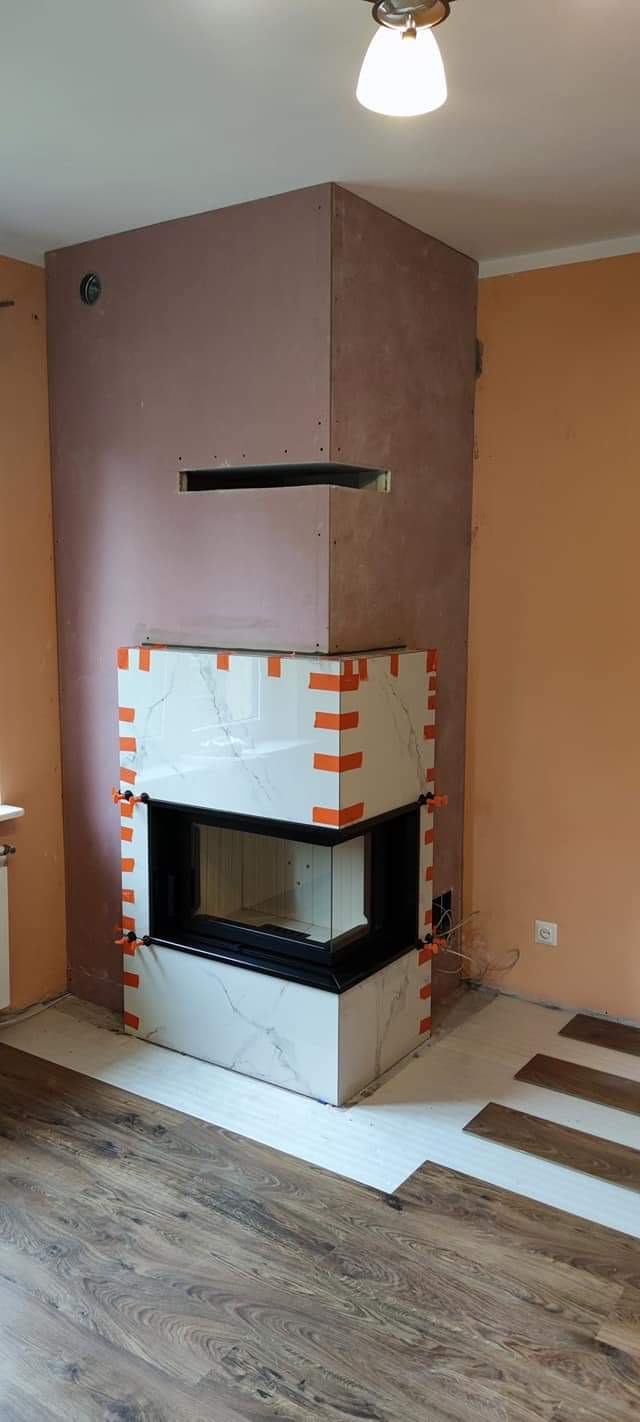 Wnętrze w trakcie budowy kominka z obudową z płyty gipsowej i marmurowych płytek, zabezpieczonych pomarańczową taśmą, z częściowo ułożoną podłogą z paneli.