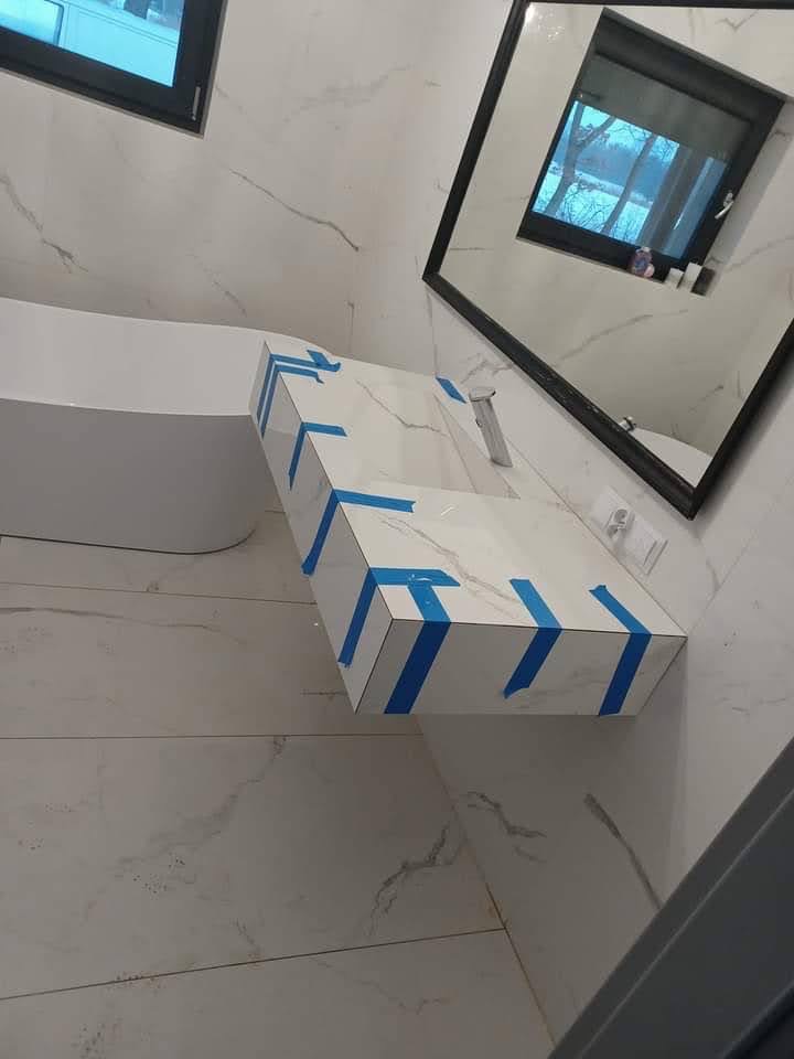 Nowoczesna łazienka w trakcie wykończenia z białą armaturą i marmurowymi płytkami, umywalka zabezpieczona niebieską taśmą malarską.