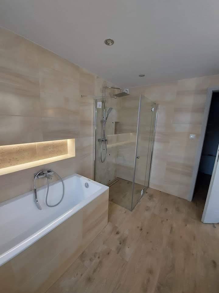 Nowoczesna łazienka z beżowymi płytkami imitującymi drewno na ścianach i podłodze, biała wanna z chromowaną baterią, przeszklona kabina prysznicowa typu walk-in z odpływem liniowym