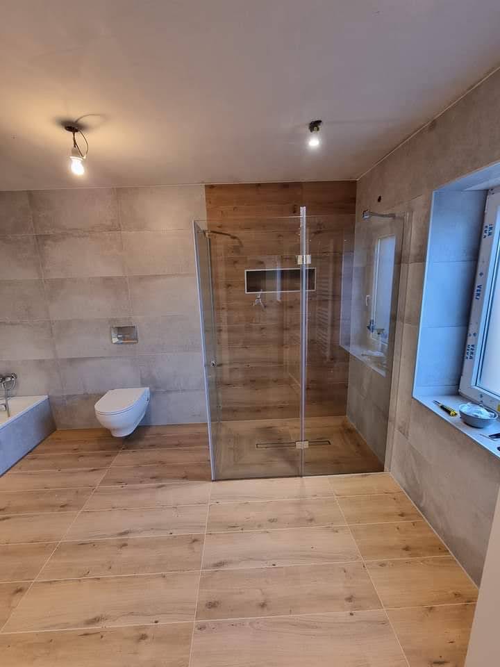 Wykończona łazienka w Szczecinie z kabiną prysznicową typu walk-in, drewnopodobną podłogą z płytek i szarą okładziną ścienną, widok na wiszącą toaletę i wannę.