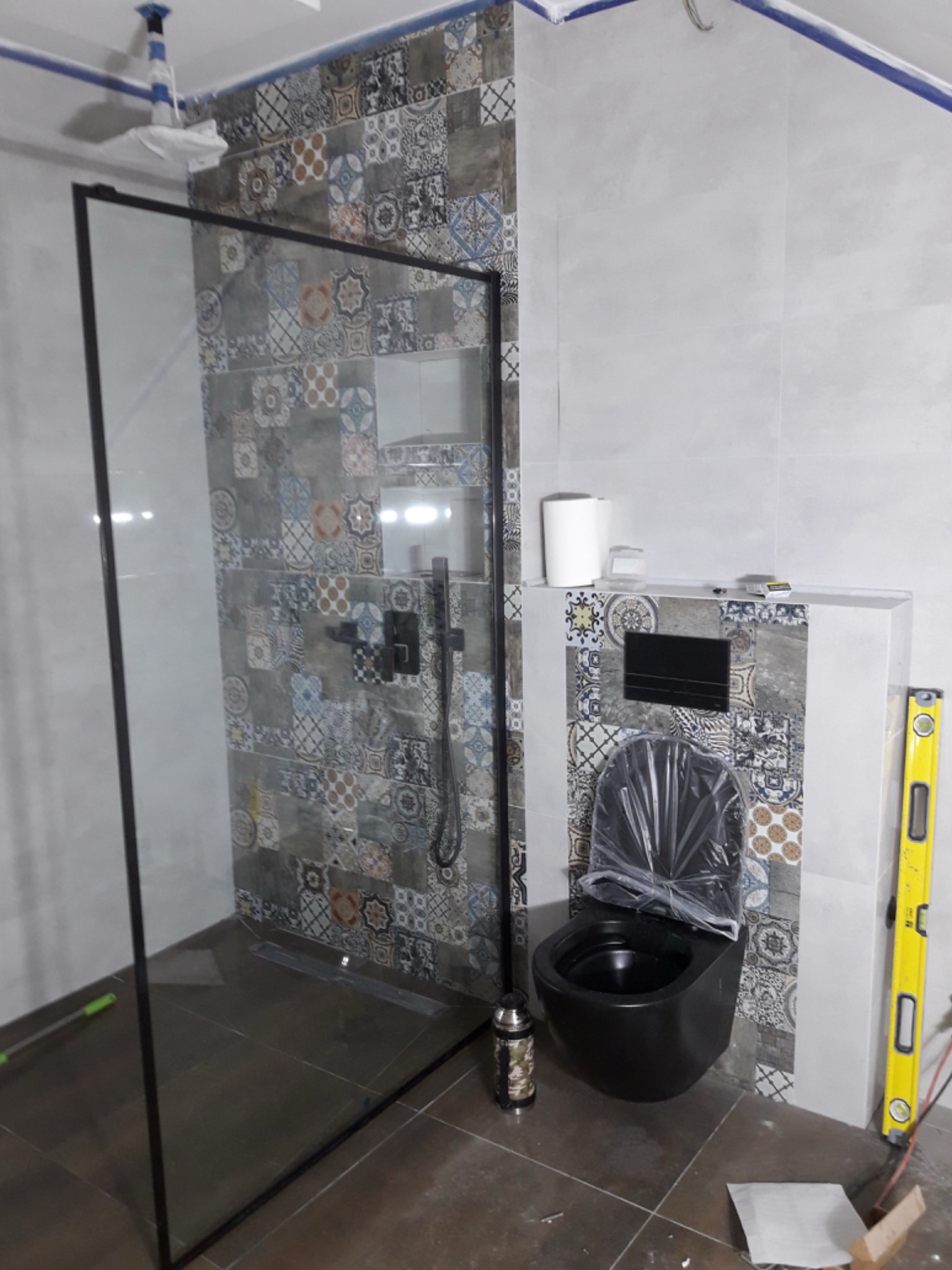 Nowoczesna łazienka w trakcie wykańczania: czarna toaleta podwieszana z folią ochronną, kabina prysznicowa typu walk-in z czarną ramą, mozaika na ścianie, poziomica oparta o ścianę, widoczne...