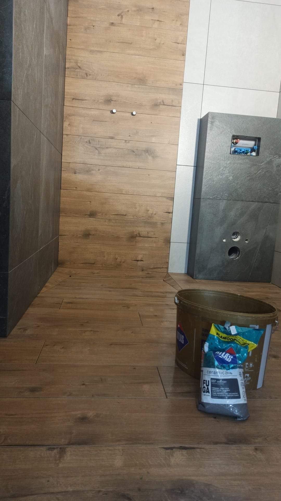 Fragment łazienki w trakcie prac wykończeniowych: widoczne płytki imitujące drewno na podłodze i ścianie, szara zabudowa WC, wiadro i worek z fugą na podłodze.