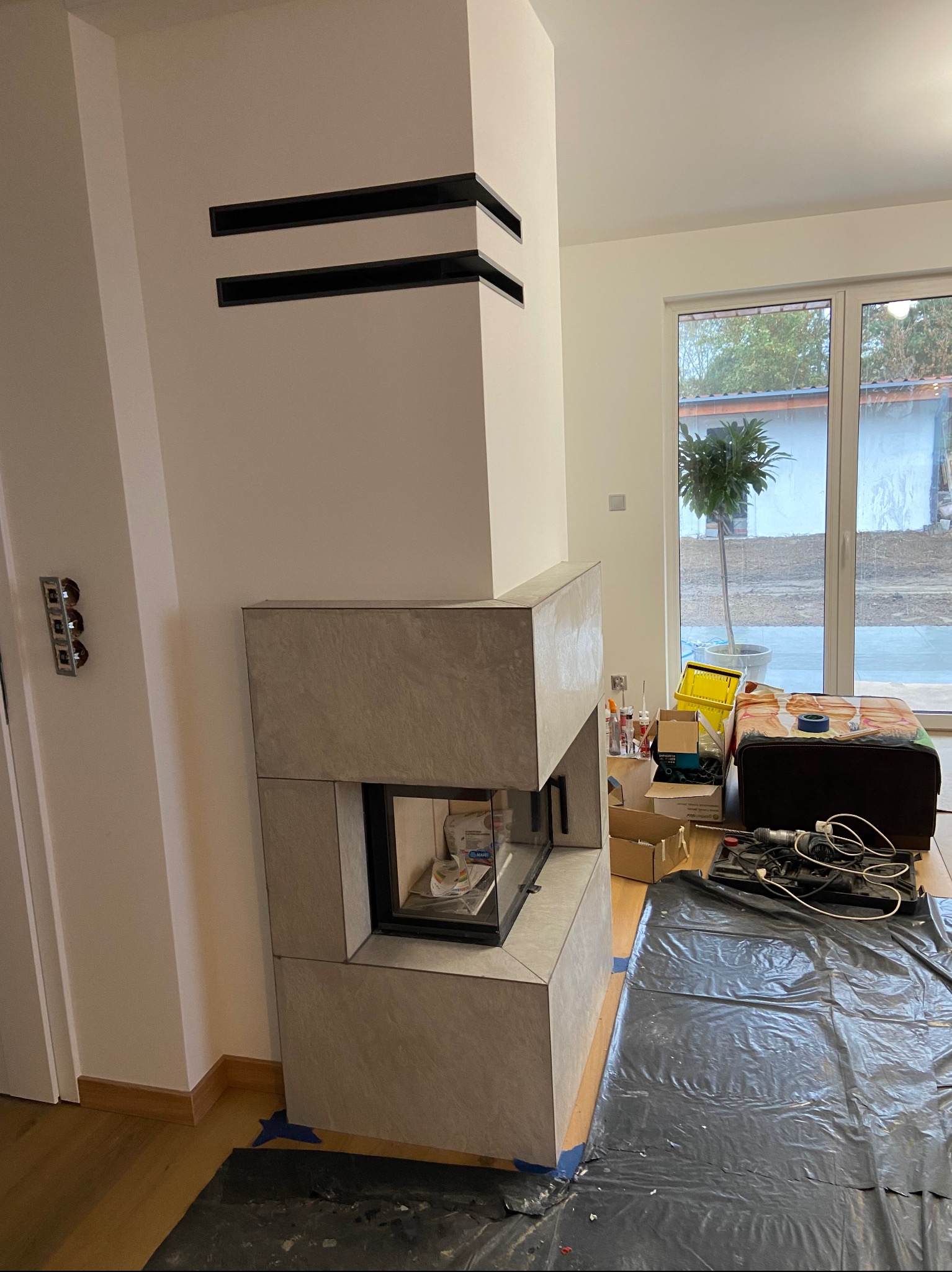 Montaż obudowy kominka z płyt kamiennych, widoczne czarne listwy dekoracyjne na ścianie, podłoga zabezpieczona folią, w tle okno tarasowe.