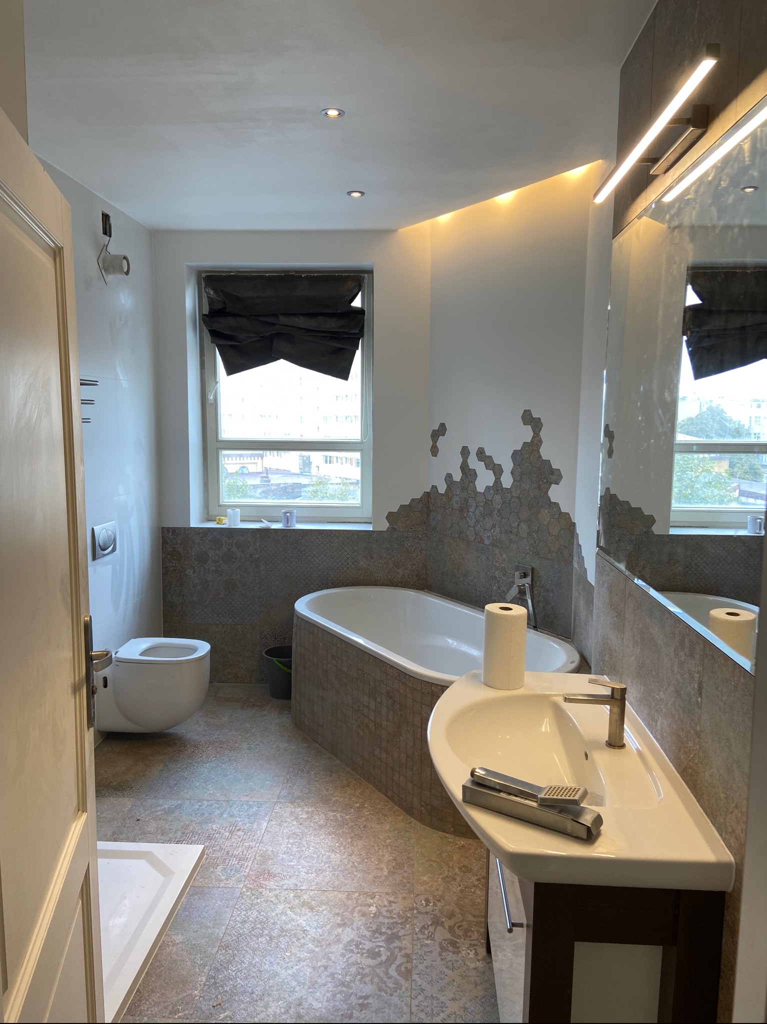 Wnętrze łazienki w trakcie wykańczania, widoczna wanna obudowana mozaiką, umywalka z baterią i prysznicem ręcznym, toaleta oraz heksagonalna mozaika na ścianie, częściowo niezakończona.