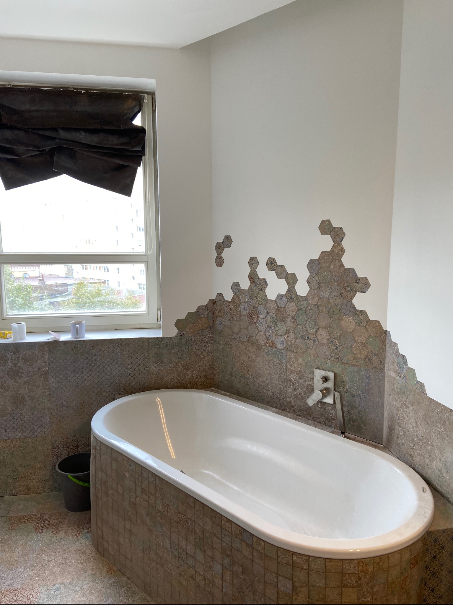 Wanna obudowana mozaiką, z fragmentem ściany wykończonym heksagonalnymi płytkami o nieregularnym kształcie, widoczny fragment okna z zasłoną rzymską, oraz akcesoria łazienkowe na parapecie.
