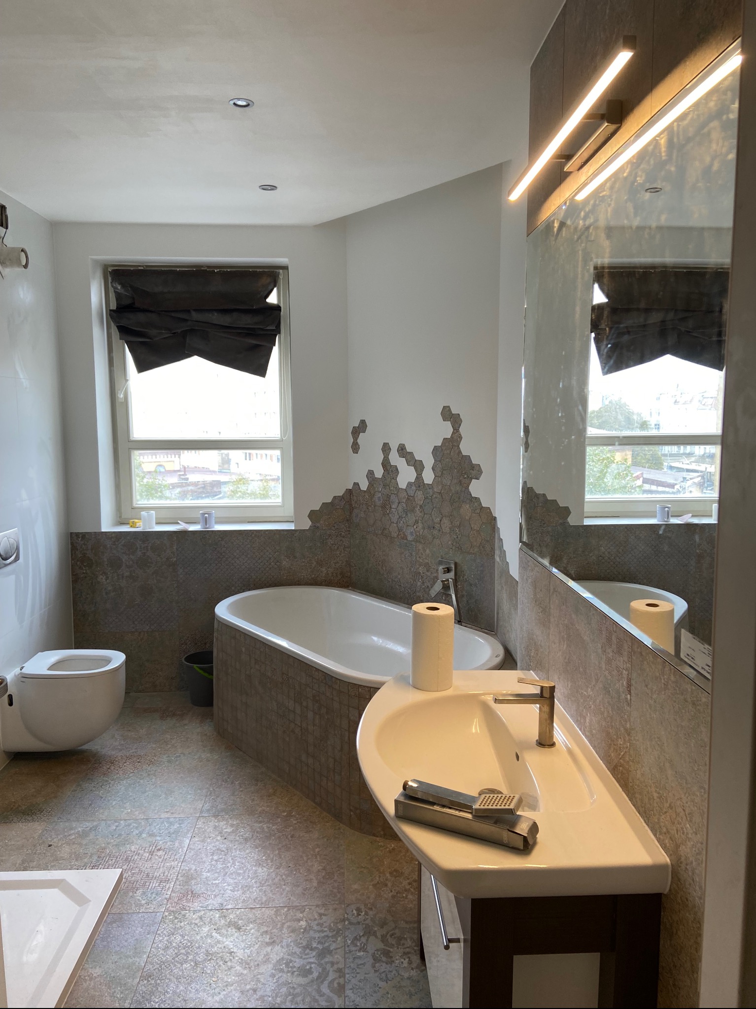 Remontowana łazienka z wanną obudowaną mozaiką, umywalką z baterią w kolorze złotym, toaletą podwieszaną i oknem z zasłoniętą roletą. Na umywalce leżą narzędzia.