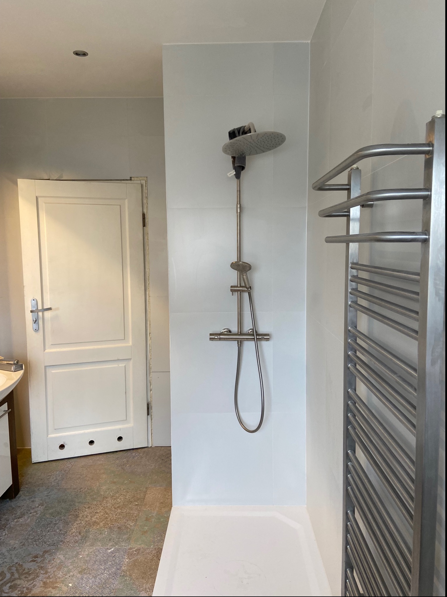 Nowoczesna kabina prysznicowa z deszczownicą i grzejnikiem drabinkowym w łazience z białymi płytkami i drzwiami z trzema otworami w dolnej części.