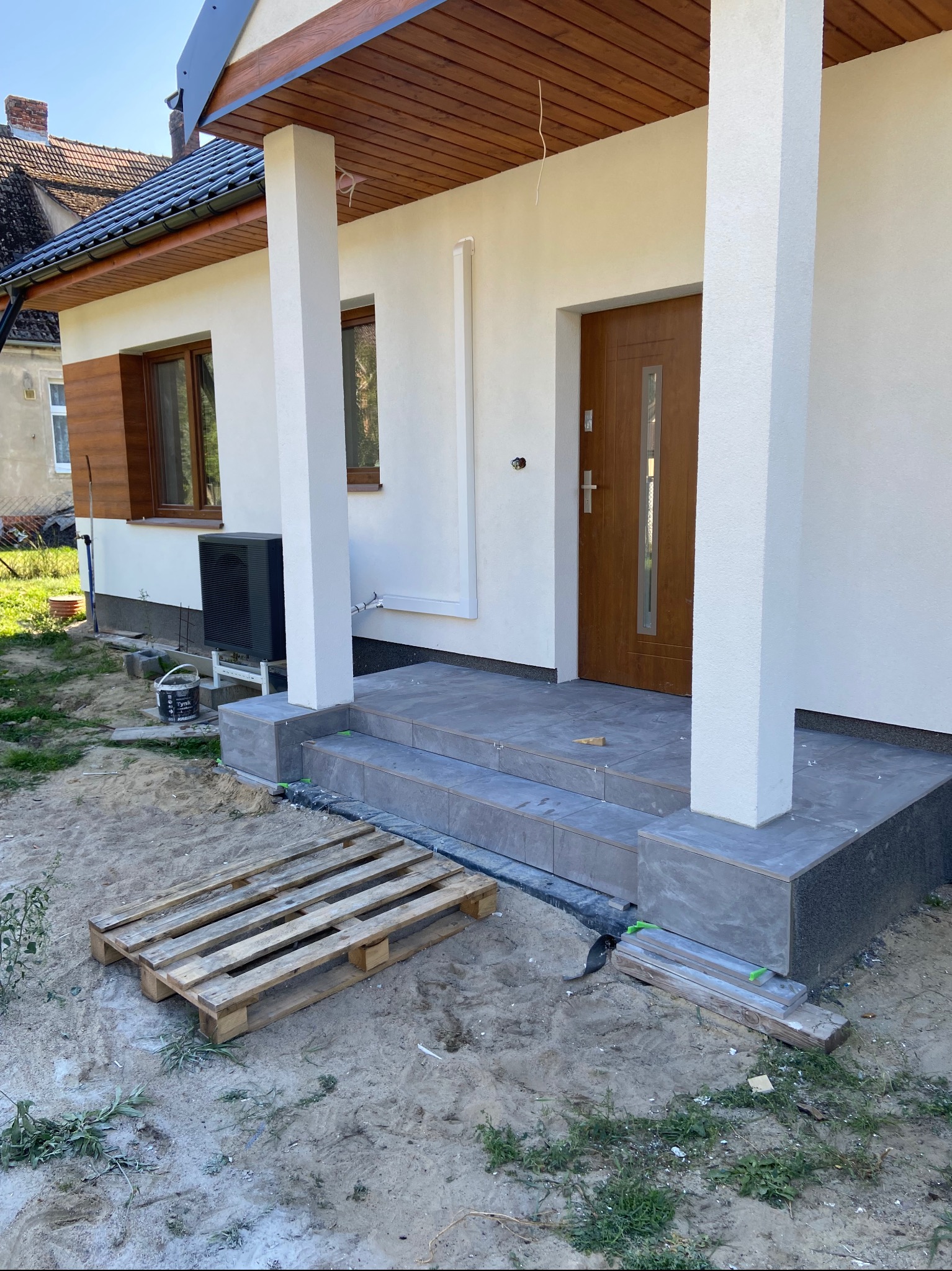 Wykończone schody wejściowe do domu z szarych płytek, drewniane drzwi wejściowe, białe kolumny i elewacja, widoczna paleta i materiały budowlane na zewnątrz.