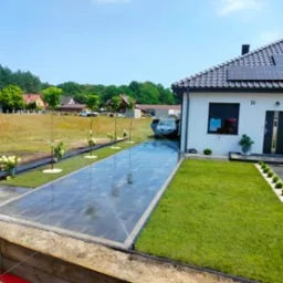 Nowo założony trawnik z rolki przylegający do błyszczącego, mokrego podjazdu z ciemnej kostki brukowej, świeżo posadzone drzewka z białym grysem u podstawy, w tle dom z panelami słonecznymi...